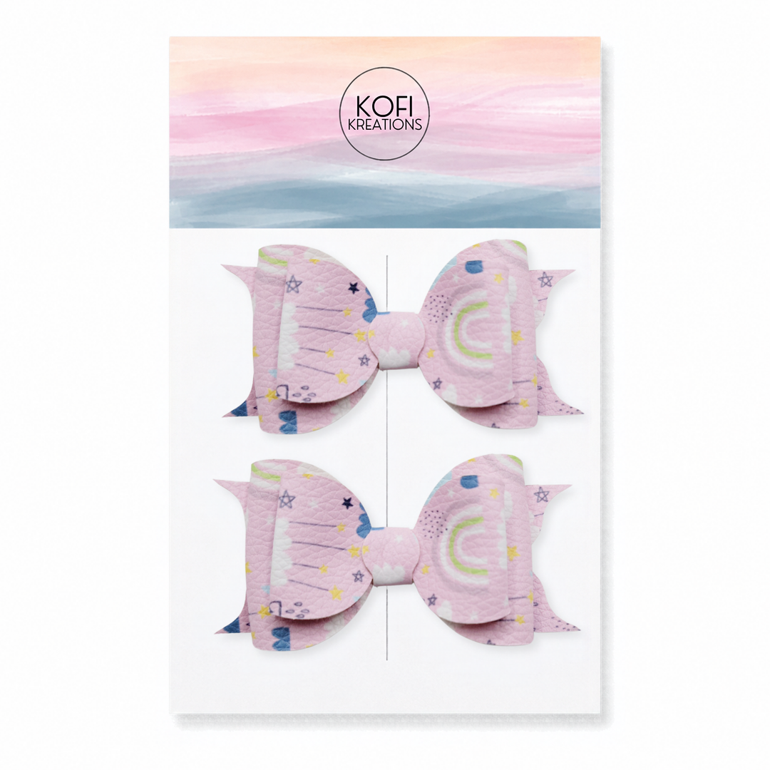 A set of two pink hair bows with celestial prints, presented on a white backing card with a pastel watercolor sky design and the brand name 'Kofi Kreations'.