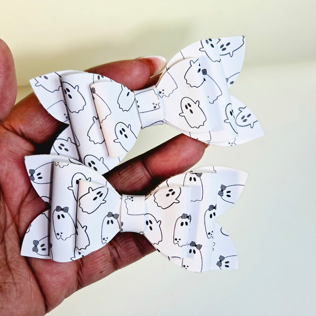 Two white faux leather hair bows with a black line-art ghost pattern, held in a person&