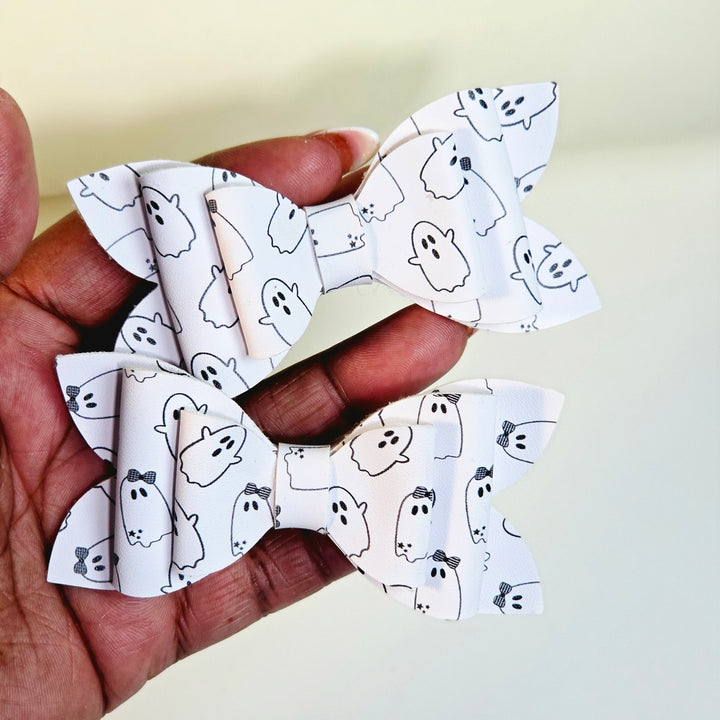 Two white faux leather hair bows with a black line-art ghost pattern, held in a person&