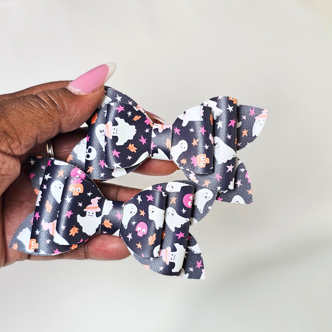 A hand with pink manicured nails holds two hair bows featuring a Halloween pattern of white ghosts, orange pumpkins, pink stars, and black stars on a dark background.