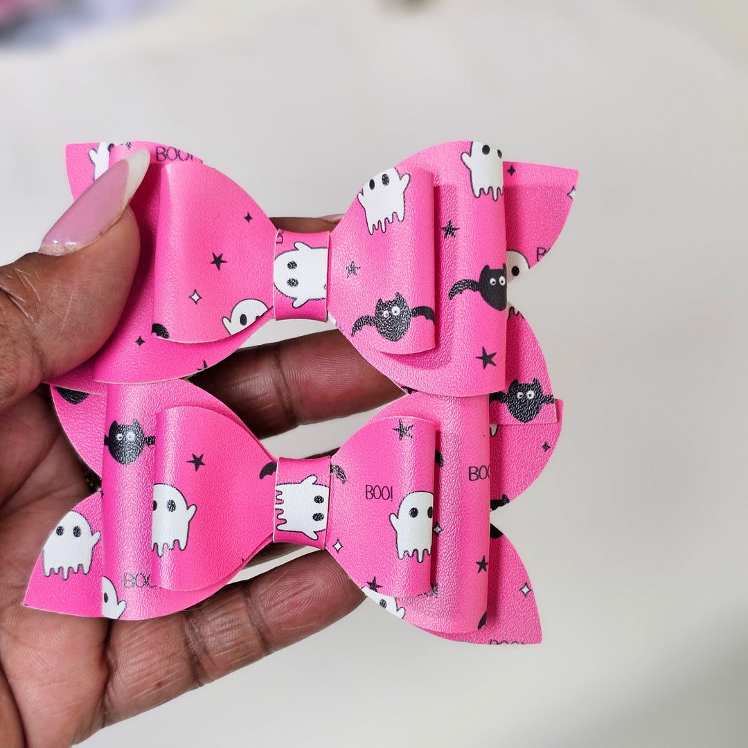 A close-up of a hand holding two bright pink hair bows decorated with white ghosts, black bats, and the word &