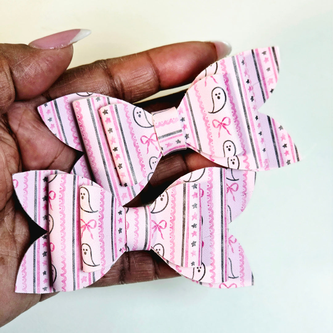 A hand holding two pink patterned hair bows with ghost and bow motifs on a light background.