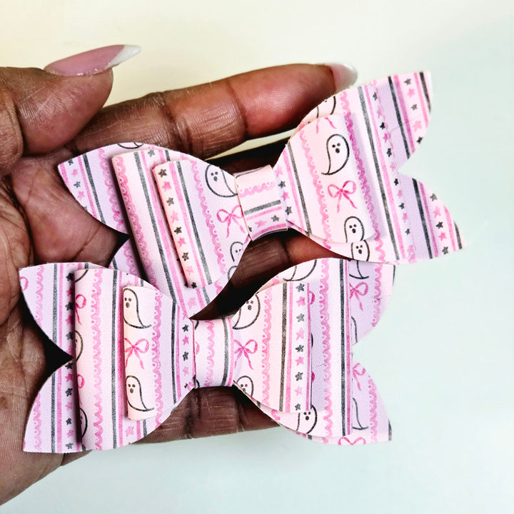 A hand holding two pink patterned hair bows with ghost and bow motifs on a light background.