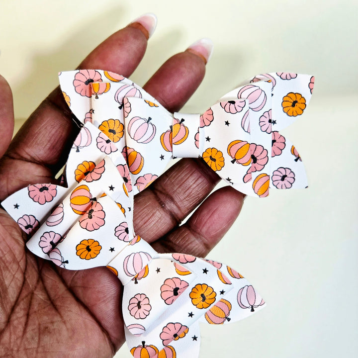 A close-up of a hand holding a white faux leather hair bow with a repeating autumn pattern of orange and pink pumpkins, striped gourds, and small black stars.