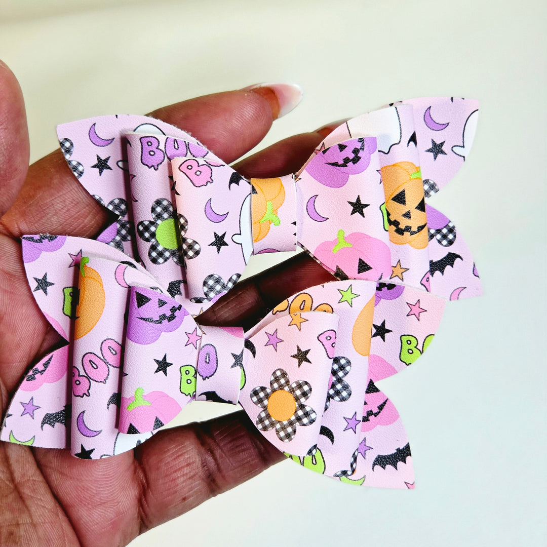 A hand holding two pink paper hair bows decorated with a Halloween pattern of pumpkins, ghosts, stars, and checkered squares.