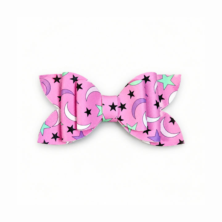 A pink faux leather hair bow with a celestial pattern featuring black stars, purple and mint green crescent moons, and white moon phases, shown on a plain white background.