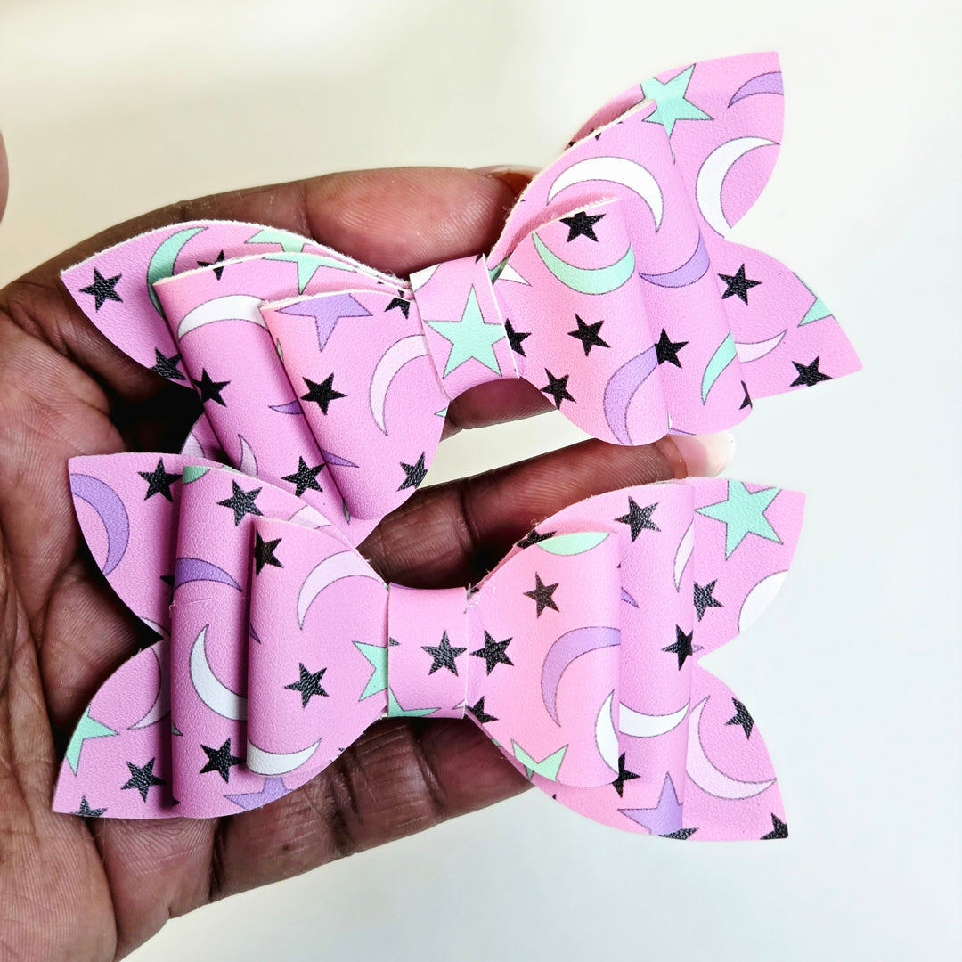 Two pink hair bows with a star and moon pattern, held in a person&