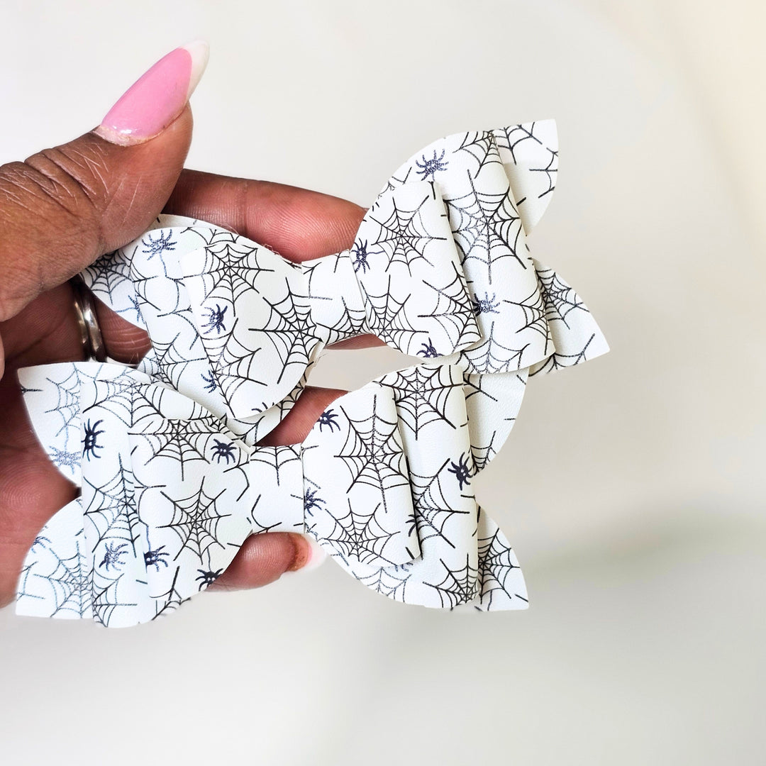 A hand with pink manicured nails holds a white faux leather hair bow decorated with a black spiderweb and spider print pattern against a plain light background.