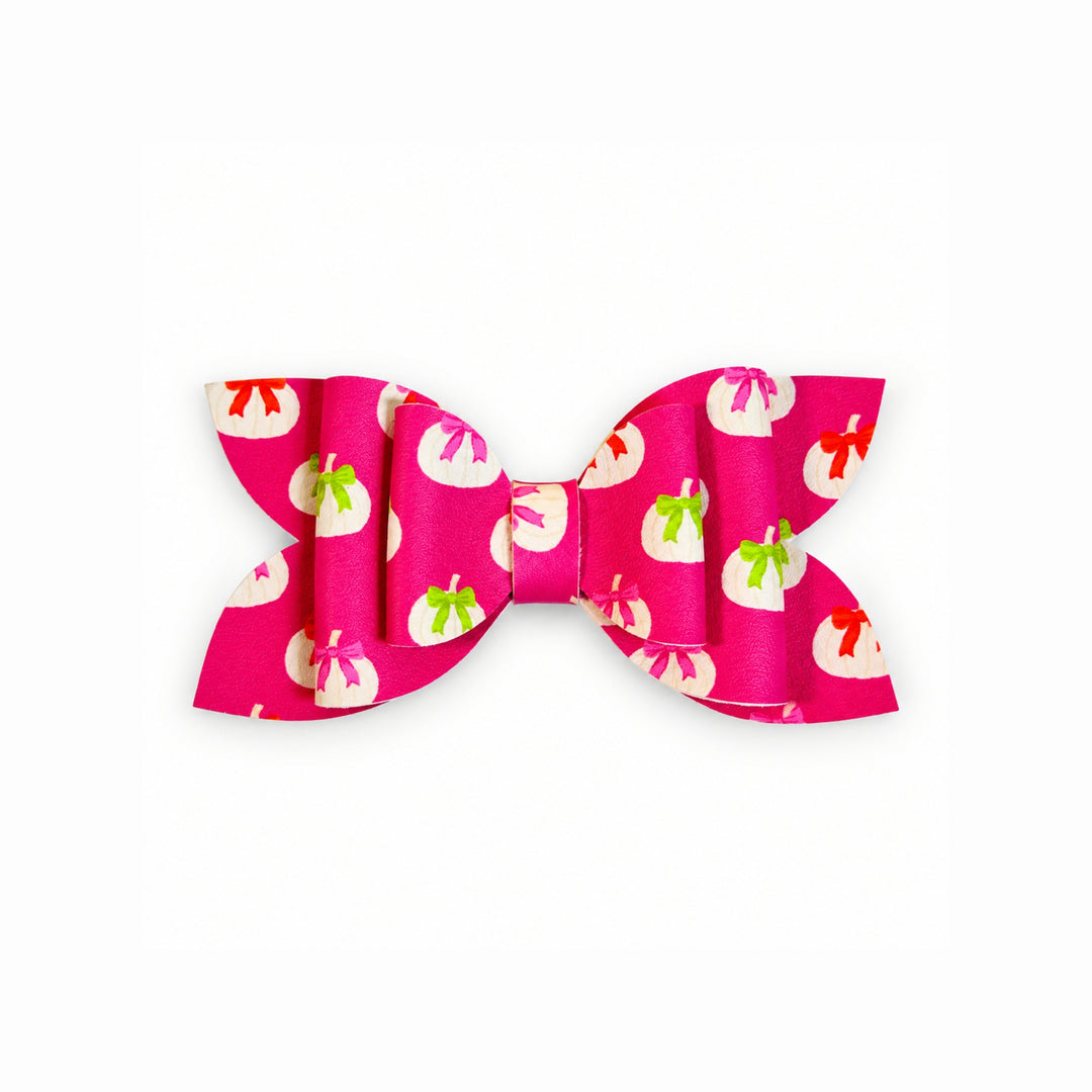 A faux leather hair bow featuring a vibrant pink background with a repeating pattern of white pumpkins decorated with red and green accents, set against a plain white background.