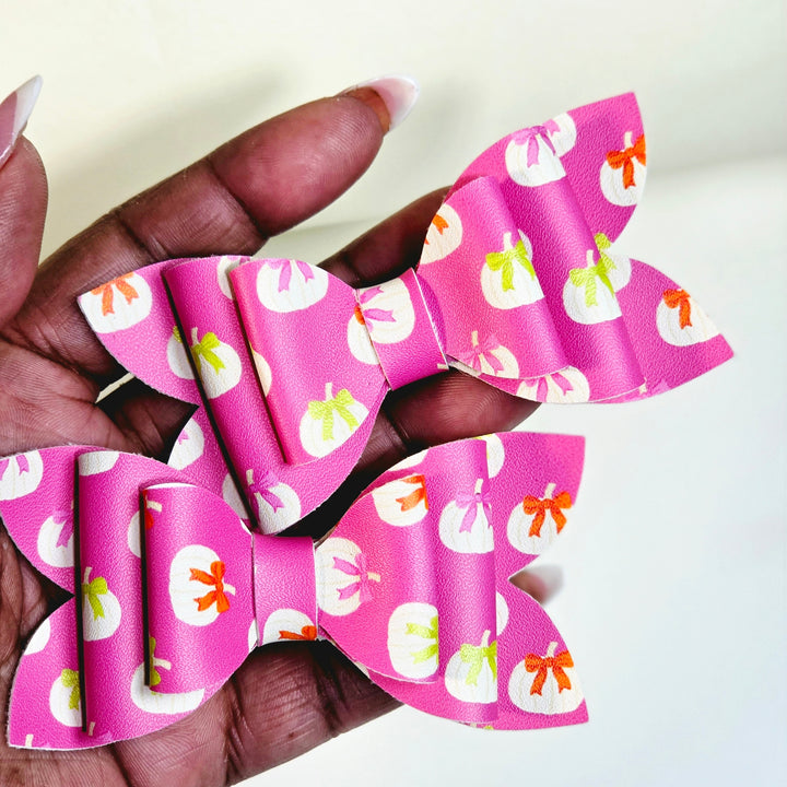 A hand holding two pink paper bow hair clips with a white, orange, and green pumpkin pattern, set against a plain light background.